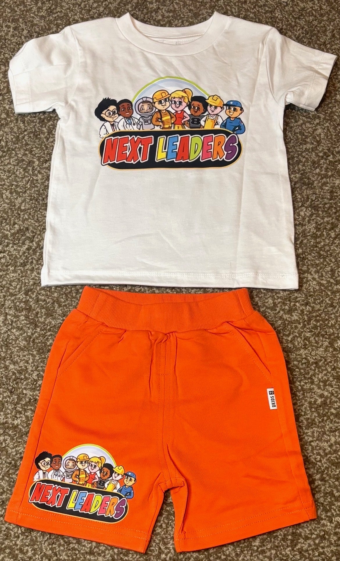 White t-shirt and orange shorts set with 'Next Leaders' graphic on a brown surface
